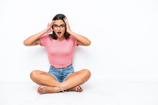 Foto joven mujer caucásica sentada en el suelo aislado sobre fondo blanco con expresión de sorpresa