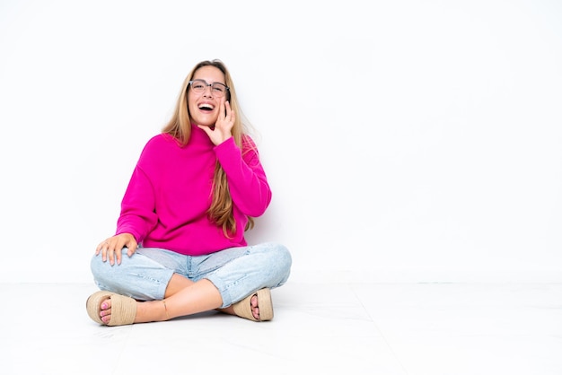 Joven mujer caucásica sentada en el suelo aislado sobre fondo blanco gritando con la boca abierta