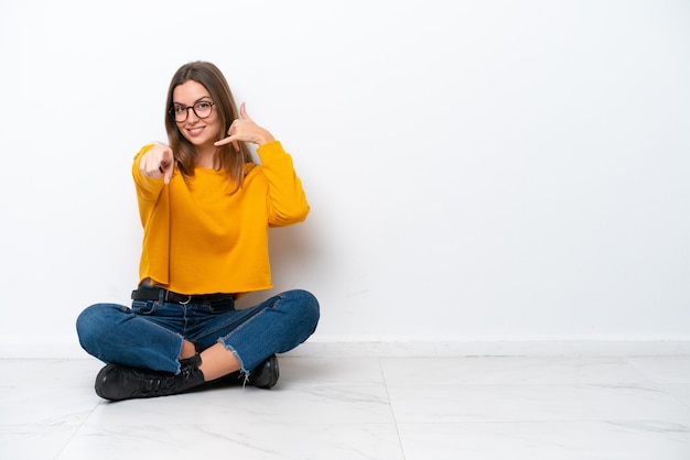 Joven mujer caucásica sentada en el suelo aislado sobre fondo blanco haciendo gesto de teléfono y apuntando hacia el frente