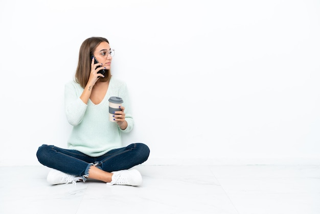 Joven mujer caucásica sentada en el suelo aislado sobre fondo blanco sosteniendo café para llevar y un móvil