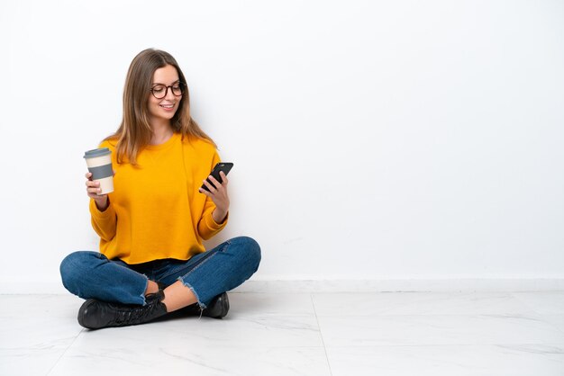 Joven mujer caucásica sentada en el suelo aislado sobre fondo blanco sosteniendo café para llevar y un móvil