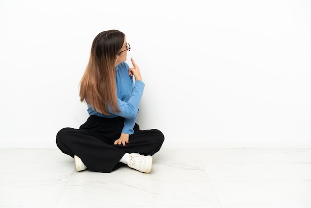 Foto joven mujer caucásica sentada en el suelo apuntando hacia atrás con el dedo índice