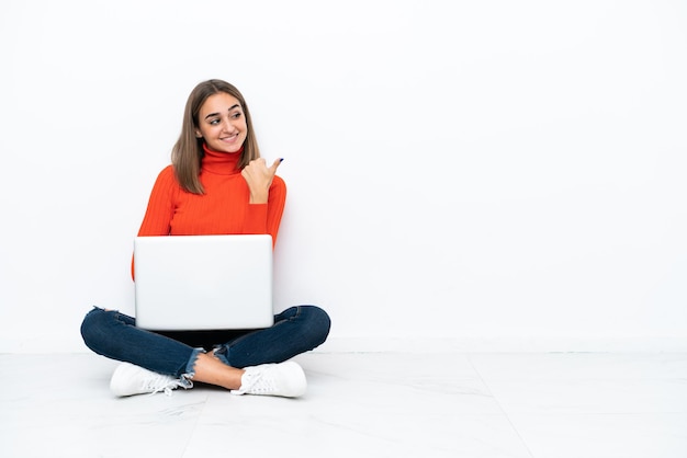 Joven mujer caucásica sentada en el suelo con una computadora portátil apuntando hacia un lado para presentar un producto