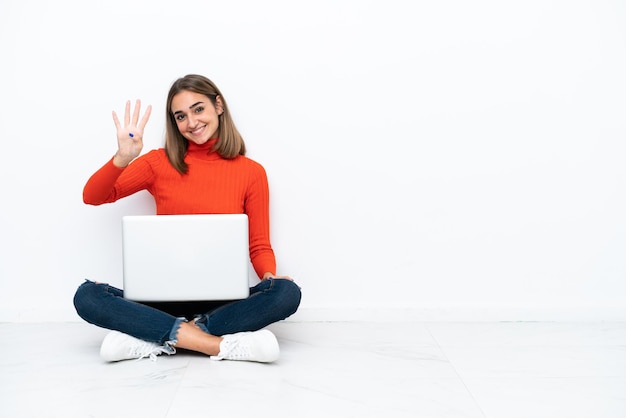 Joven mujer caucásica sentada en el suelo con una computadora portátil feliz y contando cuatro con los dedos