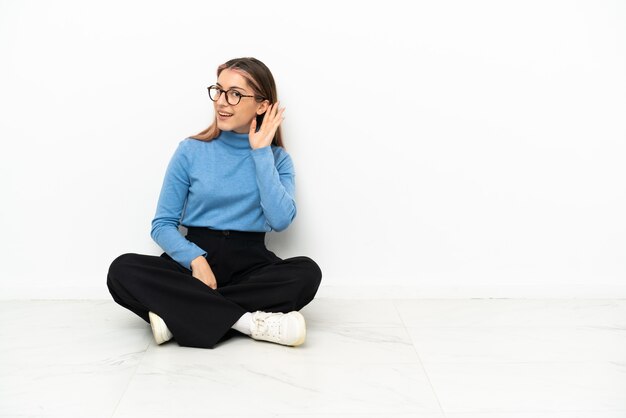 Joven mujer caucásica sentada en el suelo escuchando algo poniendo la mano en la oreja