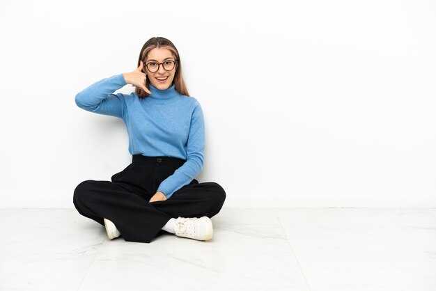 Joven mujer caucásica sentada en el suelo haciendo gesto de teléfono