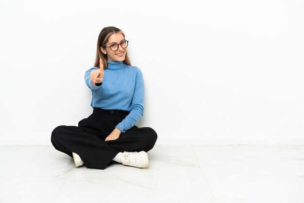 Joven mujer caucásica sentada en el suelo mostrando y levantando un dedo