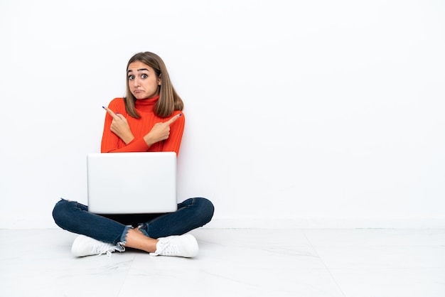 Joven mujer caucásica sentada en el suelo con un portátil apuntando a los laterales teniendo dudas