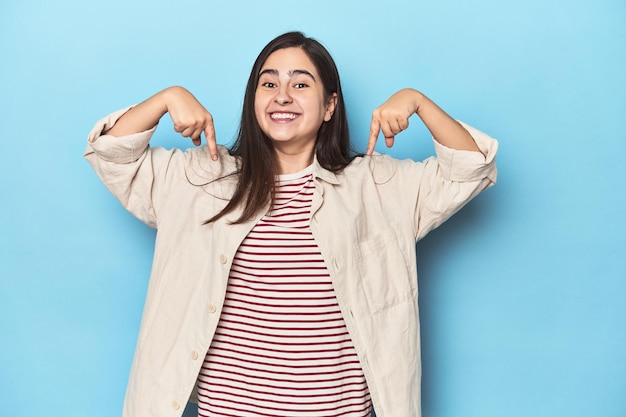 Joven mujer caucásica sobre fondo azul apunta hacia abajo con los dedos sentimiento positivo