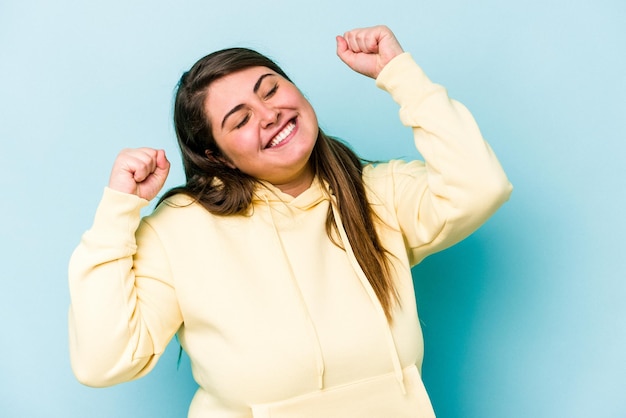 Joven mujer caucásica con sobrepeso aislada de fondo azul celebrando un día especial salta y levanta los brazos con energía