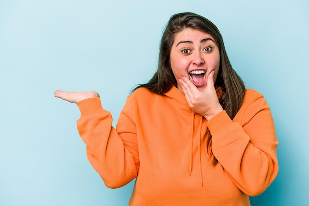 Joven mujer caucásica con sobrepeso aislada de fondo azul tiene espacio para copiar en una palma, mantenga la mano sobre la mejilla Asombrada y encantada