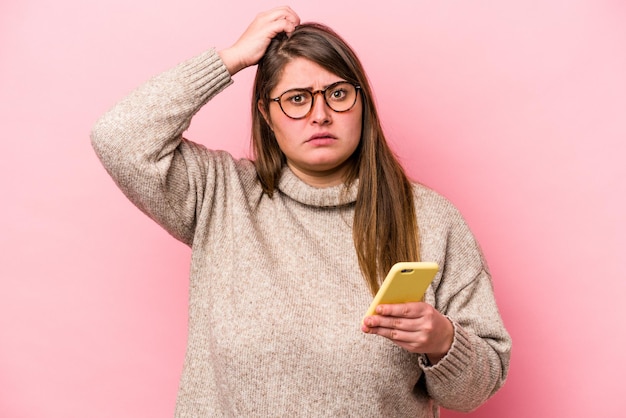 Joven mujer caucásica con sobrepeso que sostiene un teléfono móvil aislado en un fondo rosado sorprendida, ha recordado una reunión importante.