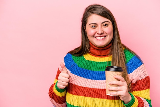 Joven mujer caucásica con sobrepeso sosteniendo café para llevar aislado sobre fondo rosa sonriendo y levantando el pulgar