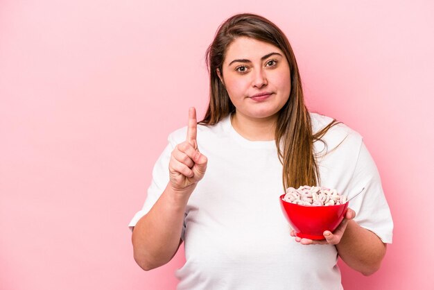 Joven mujer caucásica con sobrepeso sosteniendo un tazón de cereales aislado en un fondo rosado que muestra el número uno con el dedo.