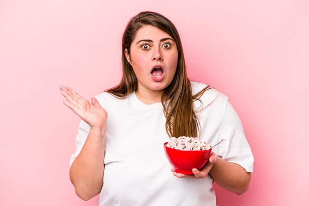 Joven mujer caucásica con sobrepeso sosteniendo un tazón de cereales aislado en un fondo rosado sorprendida y conmocionada.