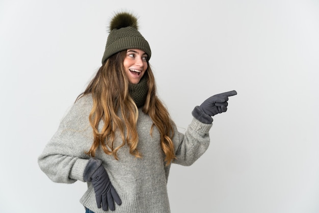 Joven mujer caucásica con sombrero de invierno aislado sobre fondo blanco apuntando con el dedo hacia el lado y presentando un producto