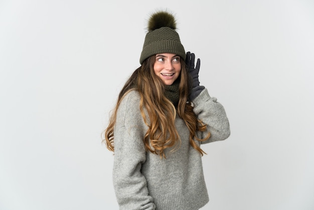 Joven mujer caucásica con sombrero de invierno aislado sobre fondo blanco escuchando algo poniendo la mano en la oreja