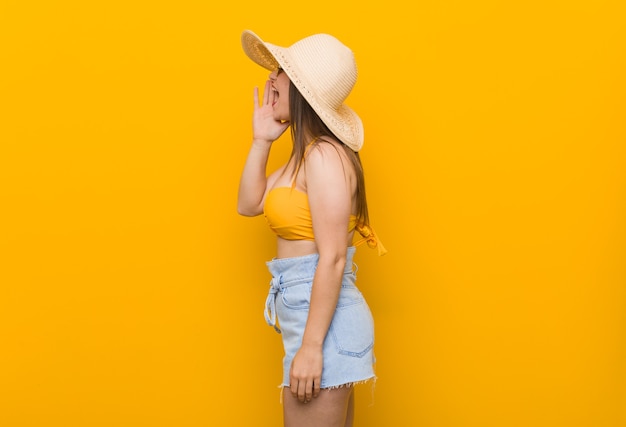 Foto joven mujer caucásica con sombrero de paja, mirada de verano gritando y sosteniendo la palma cerca de la boca abierta.