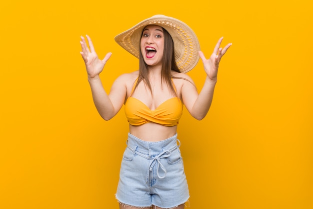 Joven mujer caucásica con un sombrero de paja, mirada de verano recibiendo una agradable sorpresa, emocionado y levantando las manos.