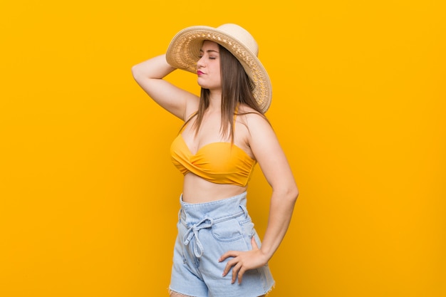 Joven mujer caucásica con un sombrero de paja, mirada de verano tocando la parte posterior de la cabeza, pensando y haciendo una elección.