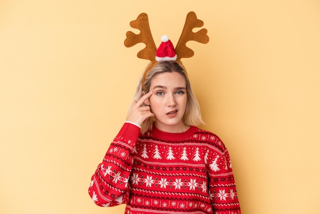 Joven mujer caucásica con un sombrero de reno de Navidad aislado sobre fondo amarillo mostrando un gesto de decepción con el dedo índice.