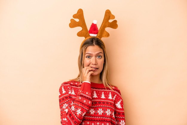 Joven mujer caucásica con un sombrero de reno de Navidad aislado sobre fondo beige mordiéndose las uñas, nervioso y muy ansioso.