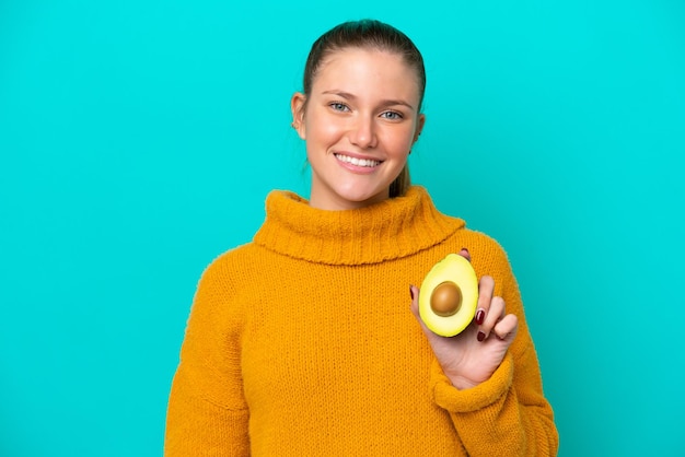 Joven mujer caucásica sosteniendo un aguacate aislado de fondo azul sonriendo mucho