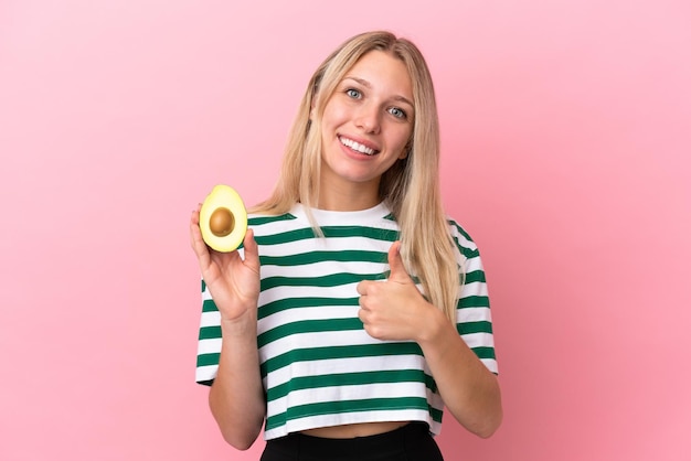 Joven mujer caucásica sosteniendo un aguacate aislado de fondo rosa dando un gesto de aprobación