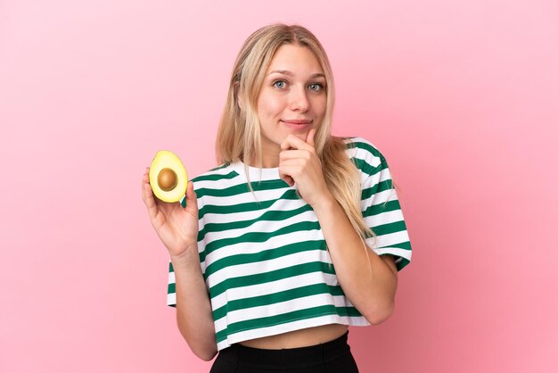 Joven mujer caucásica sosteniendo un aguacate aislado sobre fondo rosa pensando