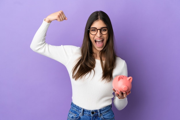 Foto joven mujer caucásica sosteniendo una alcancía aislada sobre fondo púrpura haciendo gesto fuerte