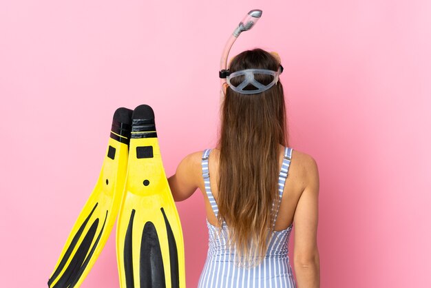 Joven mujer caucásica sosteniendo aletas y gafas de buceo aisladas sobre fondo rosa en posición trasera