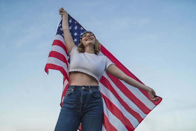 Joven mujer caucásica sosteniendo la bandera de los Estados Unidos en el cielo azul