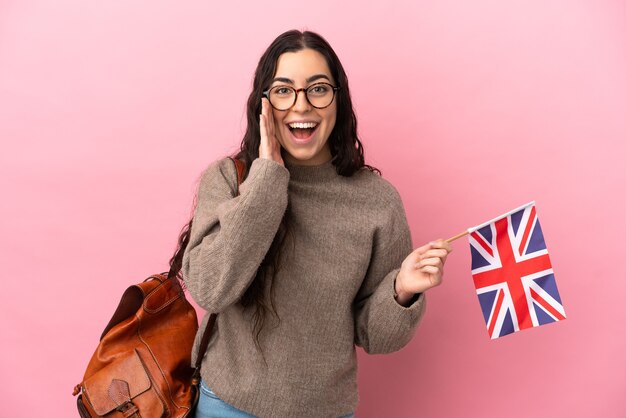 Joven mujer caucásica sosteniendo una bandera del Reino Unido aislada en la pared rosa con sorpresa y expresión facial conmocionada