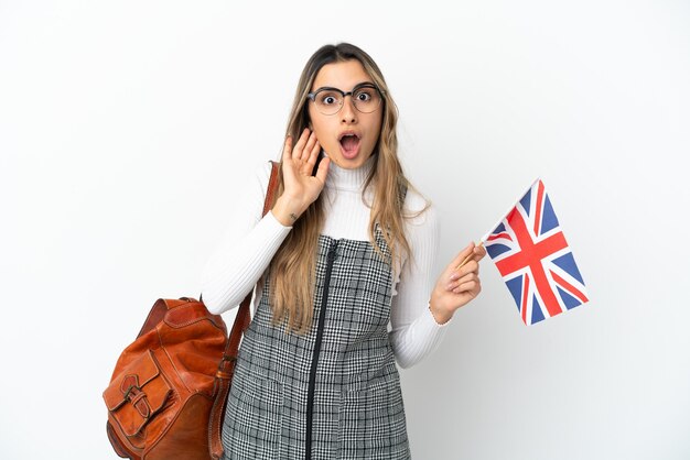 Joven mujer caucásica sosteniendo una bandera del Reino Unido aislada sobre fondo blanco con sorpresa y expresión facial conmocionada