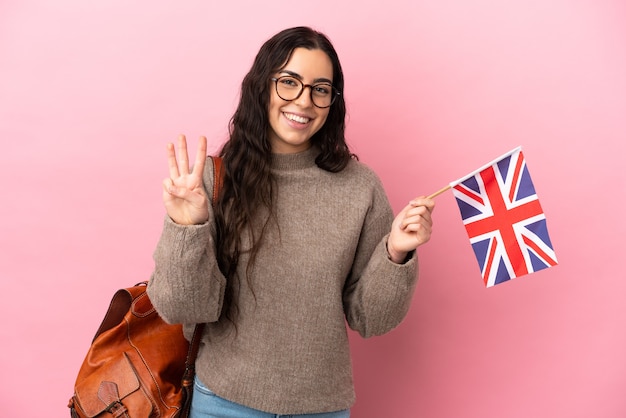 Joven mujer caucásica sosteniendo una bandera del Reino Unido aislada sobre fondo rosa feliz y contando tres con los dedos