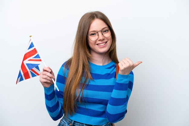 Joven mujer caucásica sosteniendo una bandera del Reino Unido aislado sobre fondo blanco apuntando hacia el lado para presentar un producto