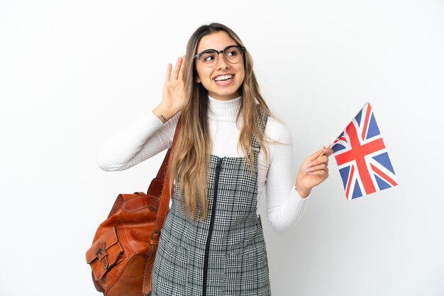 Joven mujer caucásica sosteniendo una bandera del Reino Unido aislado sobre fondo blanco escuchando algo poniendo la mano en la oreja