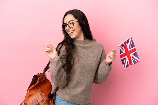 Joven mujer caucásica sosteniendo una bandera del Reino Unido aislado sobre fondo rosa dedo señalador hacia el lado y presentando un producto