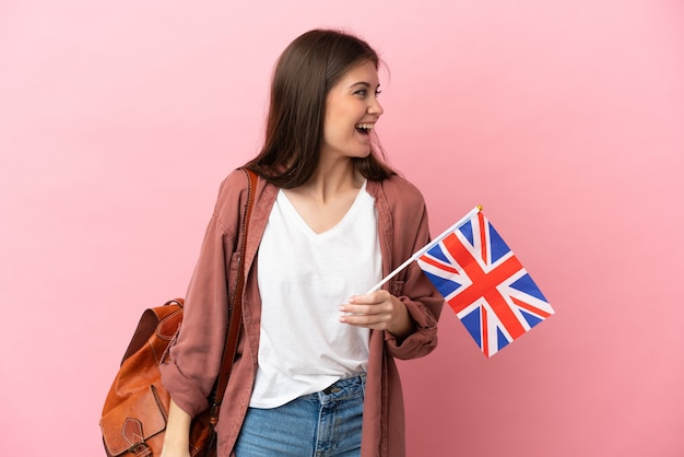 Joven mujer caucásica sosteniendo una bandera del Reino Unido aislado sobre fondo rosa riendo en posición lateral