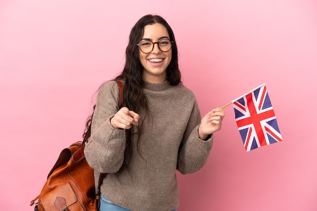 Joven mujer caucásica sosteniendo una bandera del Reino Unido aislado sobre fondo rosa sorprendido y apuntando hacia el frente