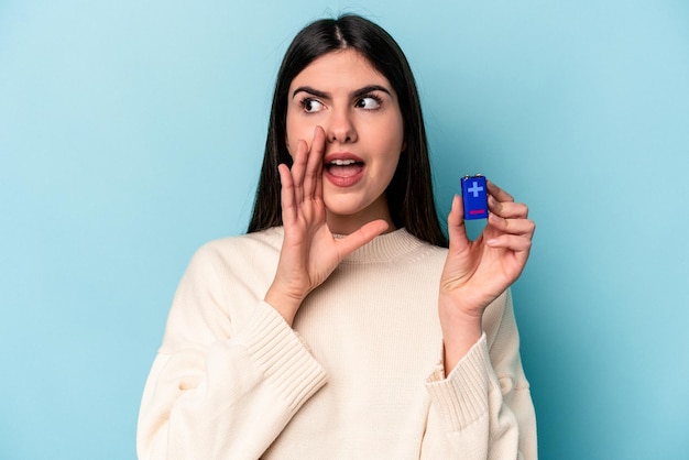 Joven mujer caucásica sosteniendo una batería aislada de fondo azul gritando y sosteniendo la palma cerca de la boca abierta