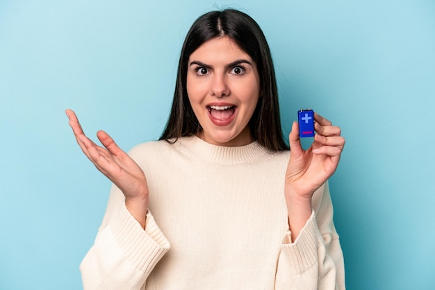Joven mujer caucásica sosteniendo una batería aislada de fondo azul recibiendo una agradable sorpresa emocionada y levantando las manos