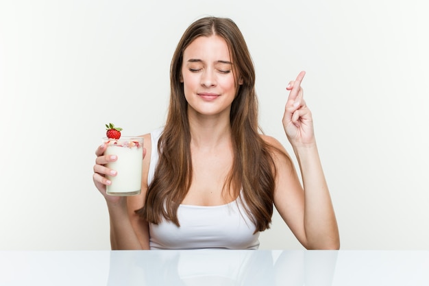 Joven mujer caucásica sosteniendo un batido cruzando los dedos para tener suerte