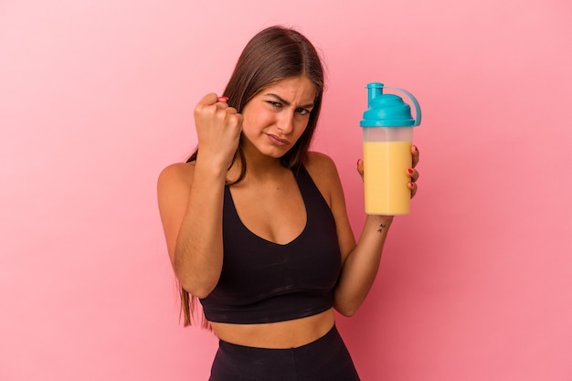 Joven mujer caucásica sosteniendo un batido de proteínas aislado sobre fondo amarillo mostrando el puño a la cámara, expresión facial agresiva.