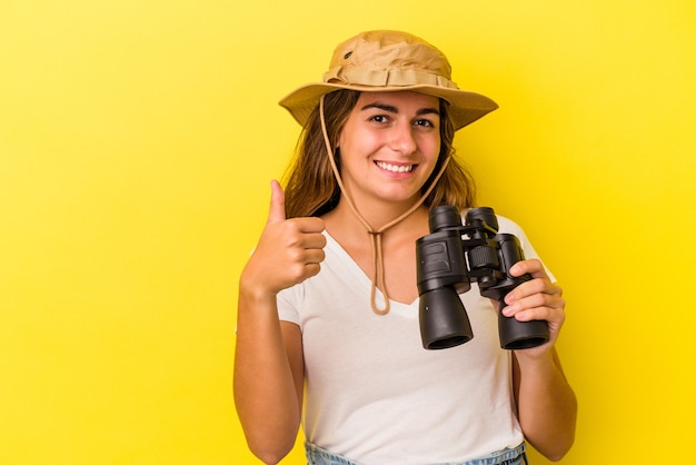 Joven mujer caucásica sosteniendo binoculares aislado sobre fondo amarillo sonriendo y levantando el pulgar hacia arriba