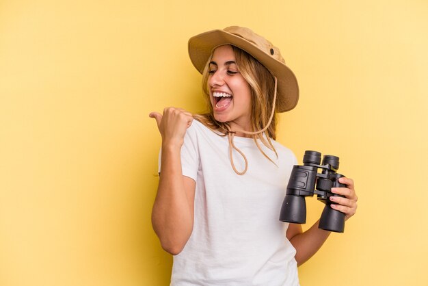 Foto joven mujer caucásica sosteniendo binoculares aislados en puntos de fondo amarillo con el dedo pulgar lejos, riendo y despreocupado.