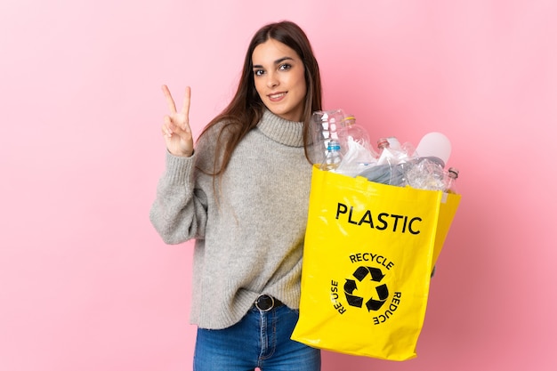 Foto joven mujer caucásica sosteniendo una bolsa llena de botellas de plástico para reciclar aislado en rosa sonriendo y mostrando el signo de la victoria