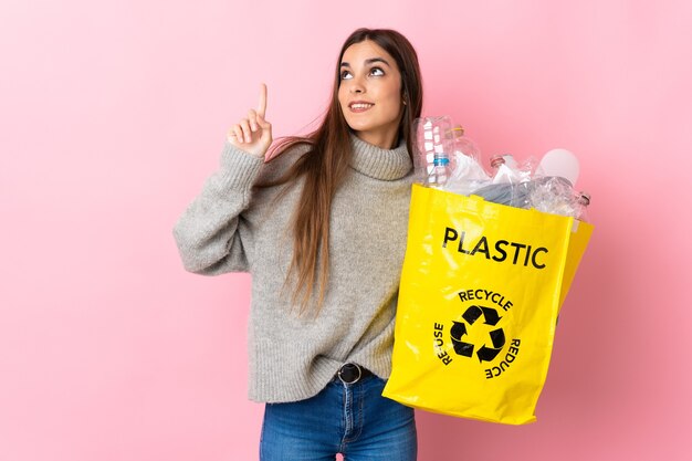 Joven mujer caucásica sosteniendo una bolsa llena de botellas de plástico para reciclar en rosa apuntando hacia una gran idea