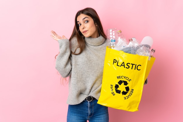 Joven mujer caucásica sosteniendo una bolsa llena de botellas de plástico para reciclar en rosa con dudas mientras levanta las manos
