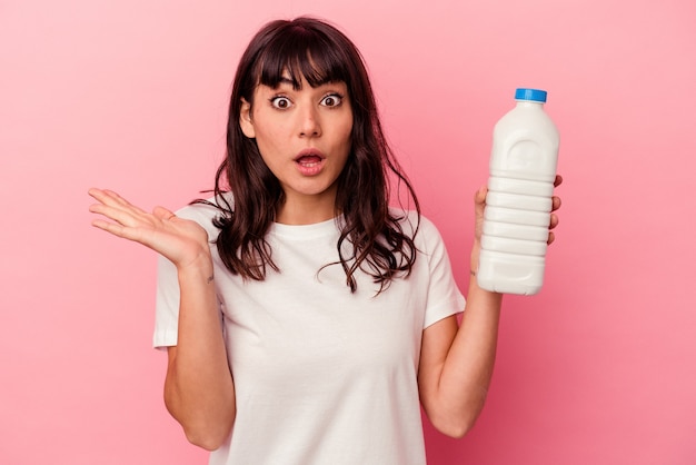 Joven mujer caucásica sosteniendo una botella de leche aislada sobre fondo rosa sorprendida y conmocionada.